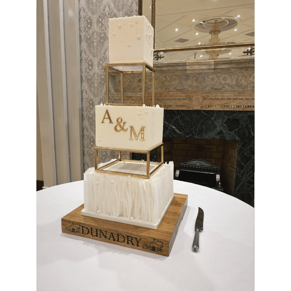 Elegant three-tier wedding cake with gold accents on a wooden base, featuring the initials "A & M" and a decorative design.