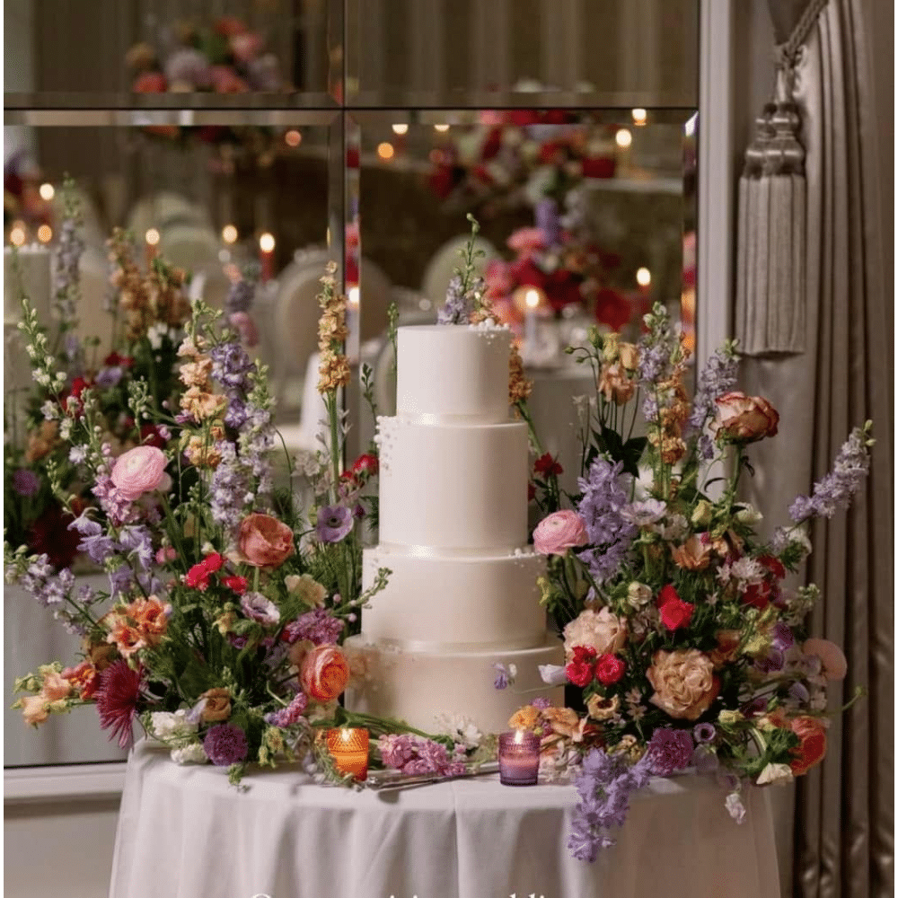 Elegant white tiered cake surrounded by colorful floral arrangements on a decorated table.