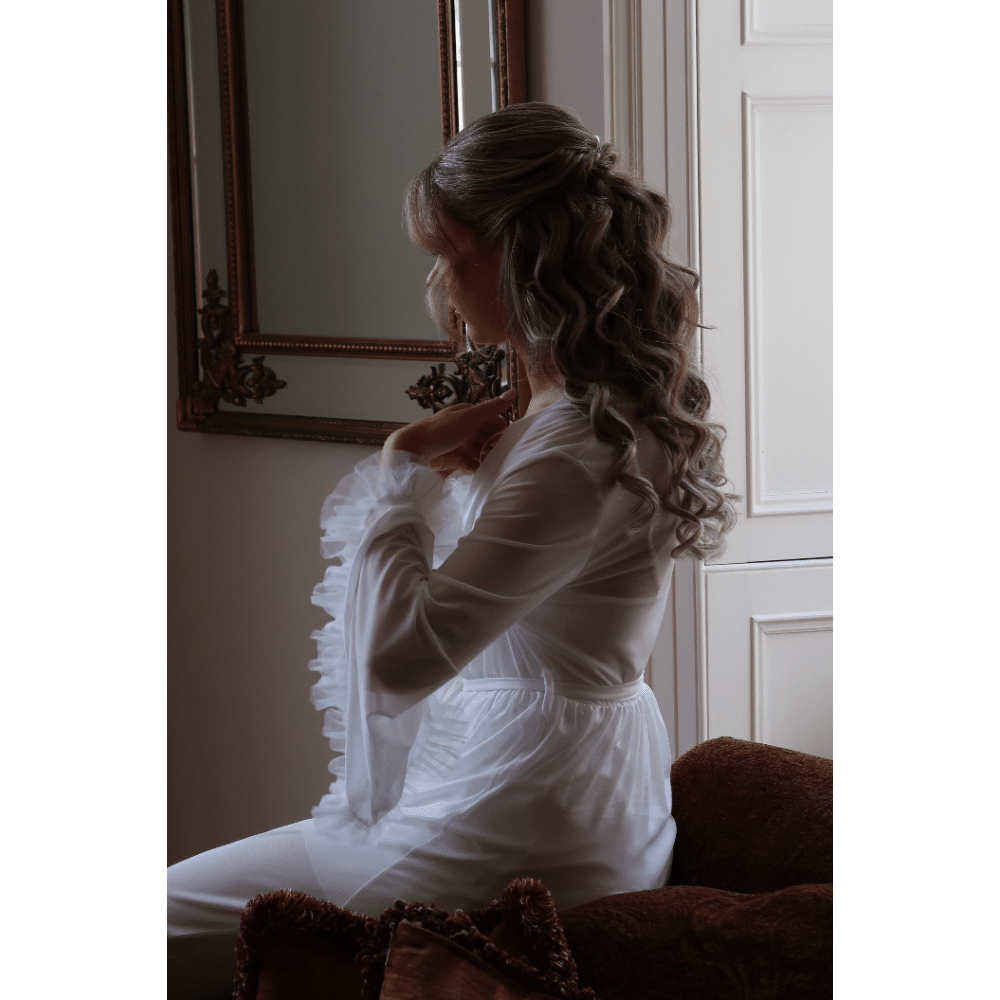 Elegant woman in white dress sitting by vintage mirror, soft lighting.