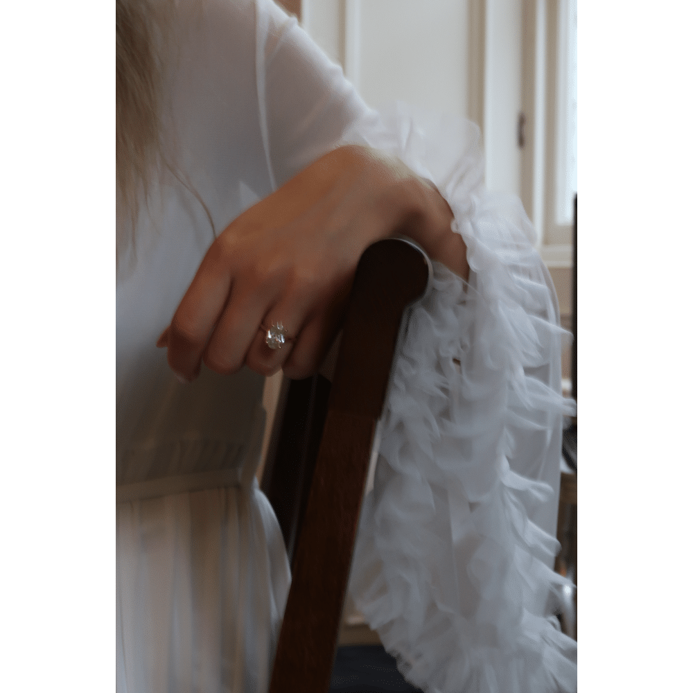 Person in white with a ruffled sleeve, wearing a ring, leaning on a chair.