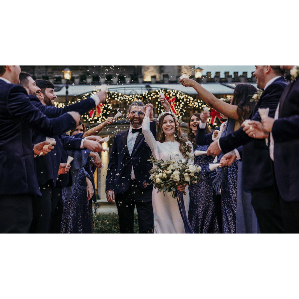 Bride and groom celebrating with guests at an outdoor wedding, surrounded by confetti and festive lights.