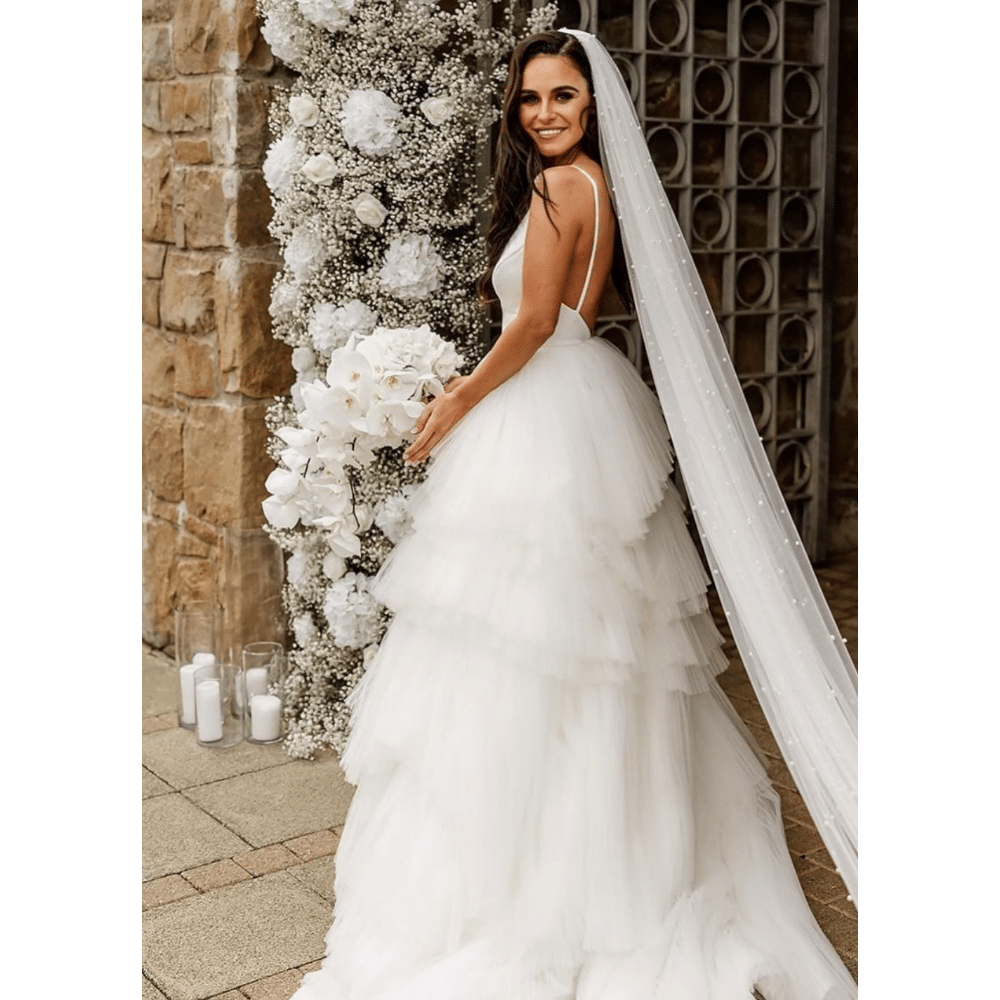 Bride in a layered white wedding dress holding a bouquet, standing beside floral decorations.