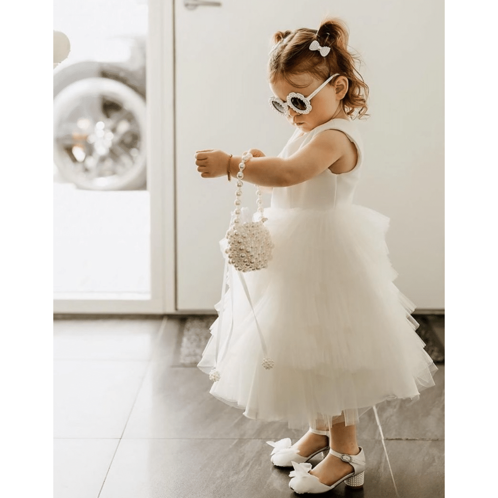 Little girl in a white dress wearing sunglasses and holding a beaded purse indoors.