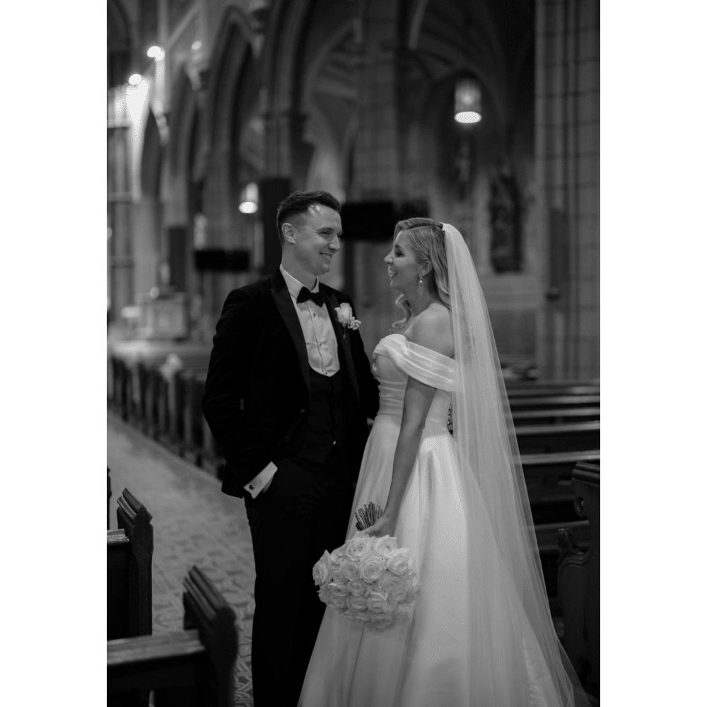 Bride and groom smiling at each other in a church, holding a bouquet.