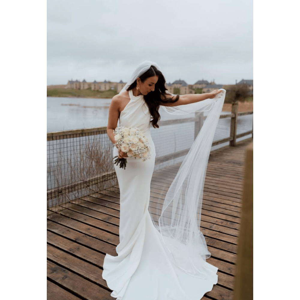 Bride in white dress holding veil and bouquet on wooden deck by a lake.