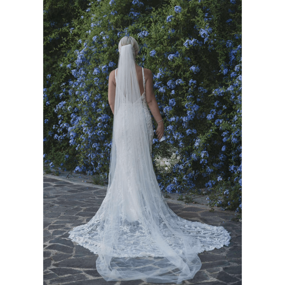 Bride in a long white gown and veil stands by a lush wall of blue flowers.