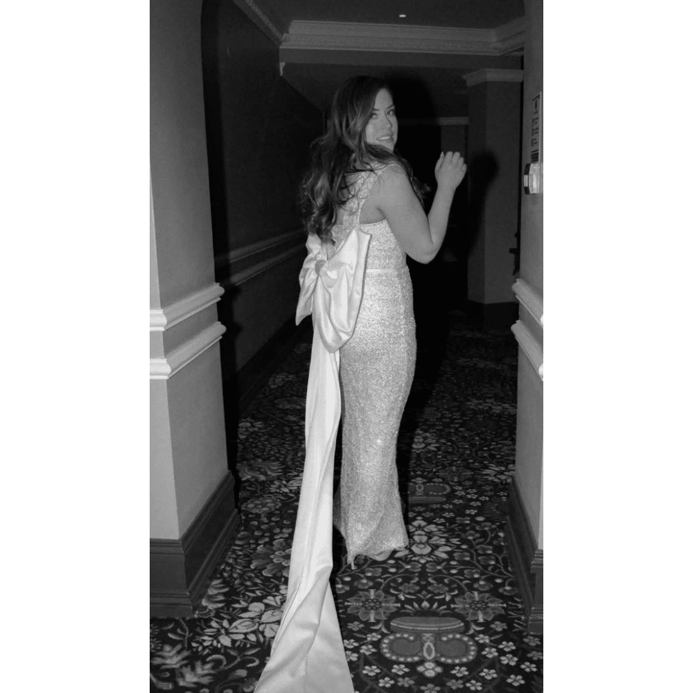 Person in a sparkling gown with a long bow sash walking down a hallway, looking back at the camera.