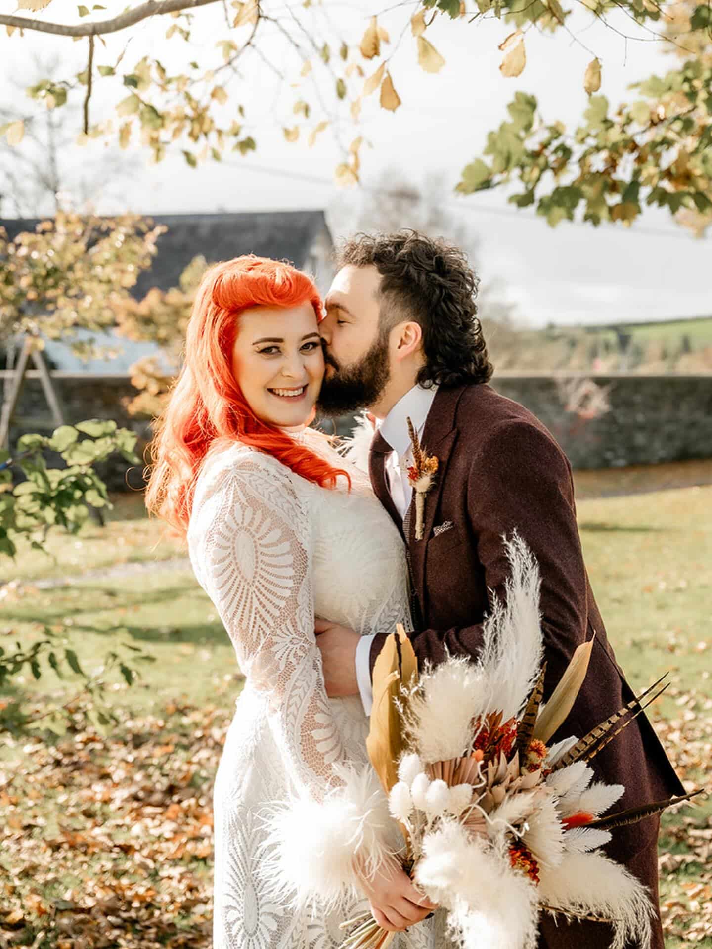 Bride and groom pose outdoors, with the groom kissing the bride's cheek. She holds a bouquet of pampas grass.