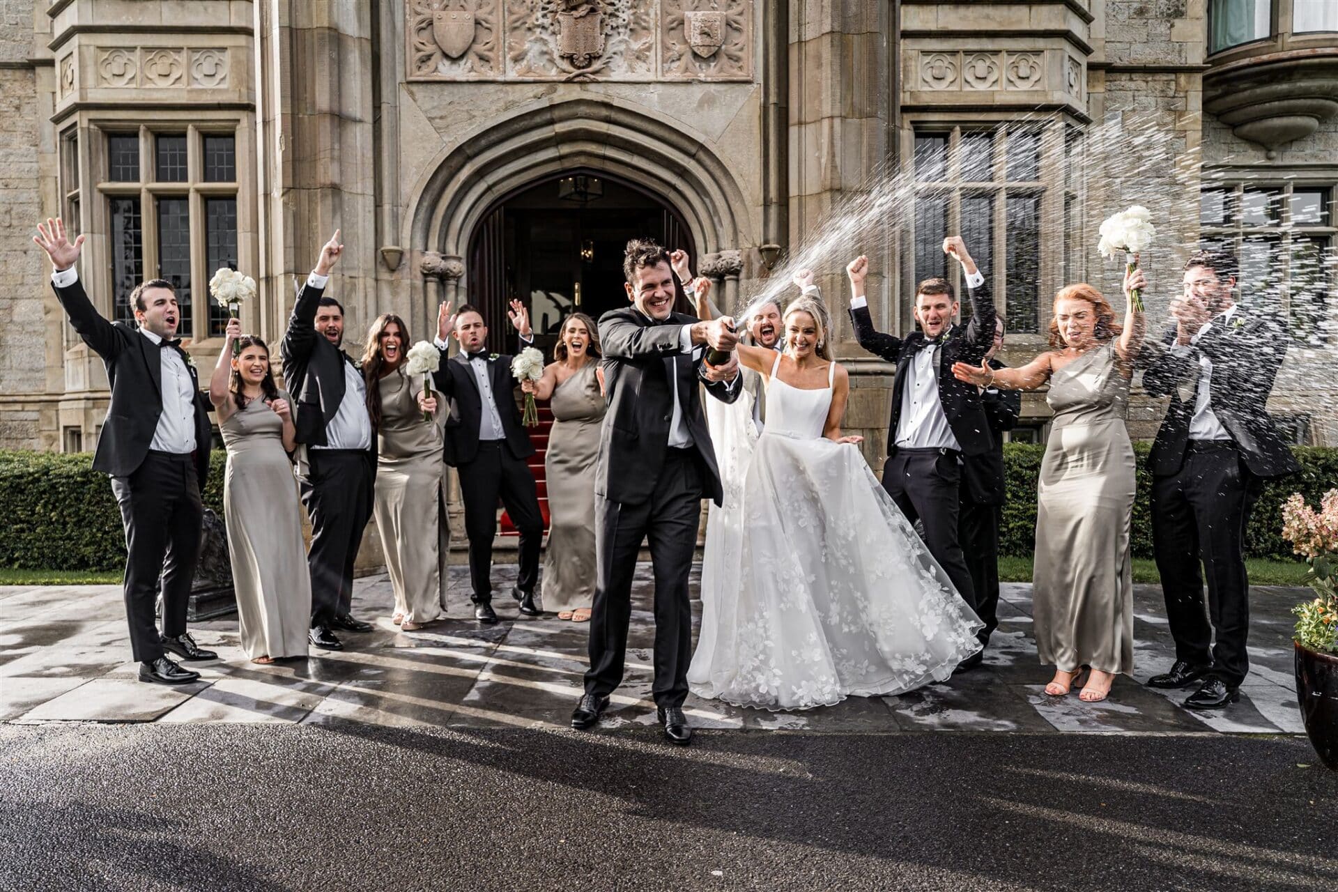 Bride and groom celebrate with bridal party, spraying champagne outside a grand building.