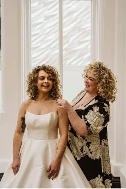 Bride in a white dress being assisted by a woman, both smiling in front of a bright window.