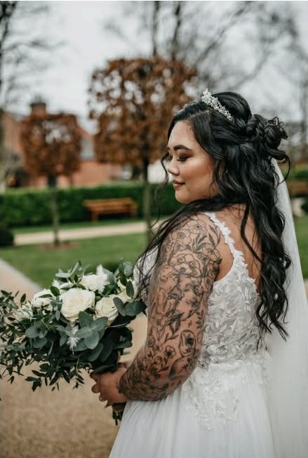 Bride with tattoos in white gown holding bouquet outdoors.