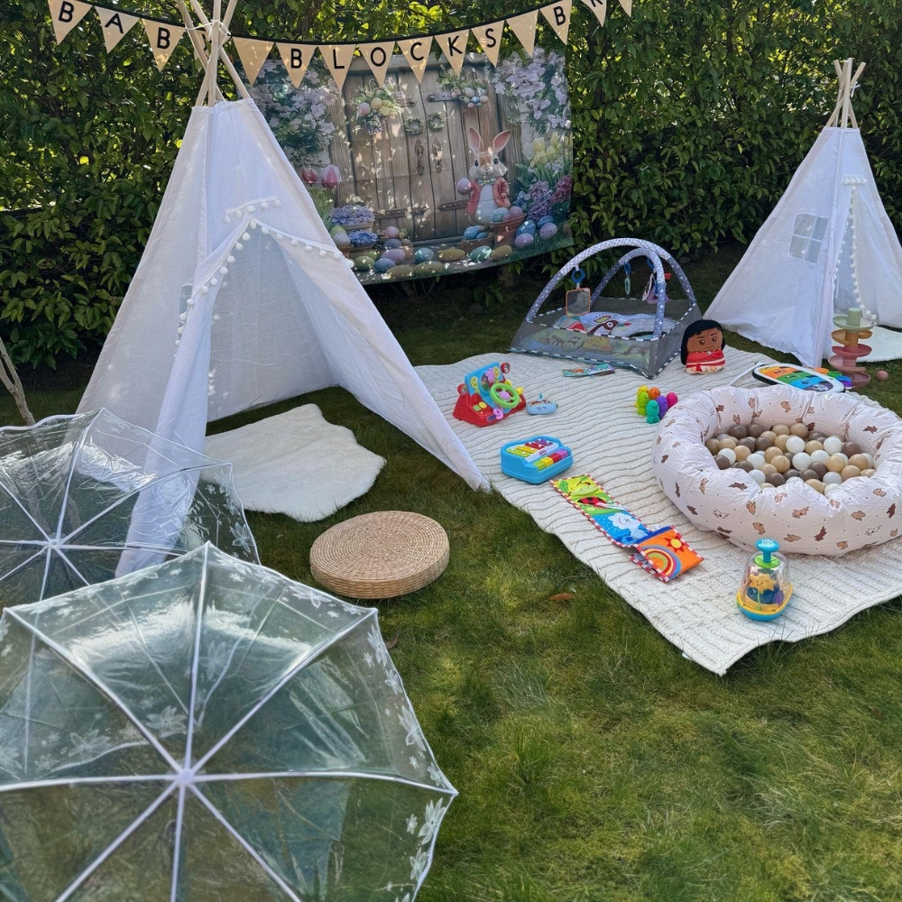 Outdoor baby play area with white teepees, toys, and umbrellas on grass.