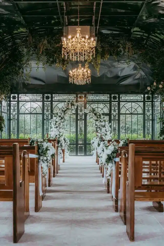 Elegant indoor wedding ceremony aisle with floral arch, chandeliers, and wooden seating.