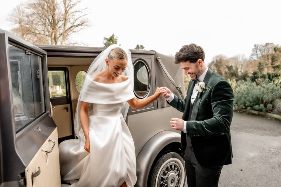 Bride stepping out of vintage car assisted by groom at Bellingham Castle.