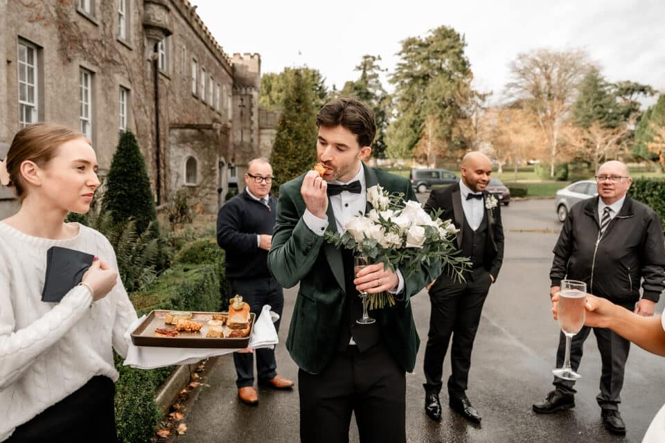 Groom in a velvet suit jacket eating canapé, holding bouquet at Bellingham Castle, surrounded by guests in formal attire.