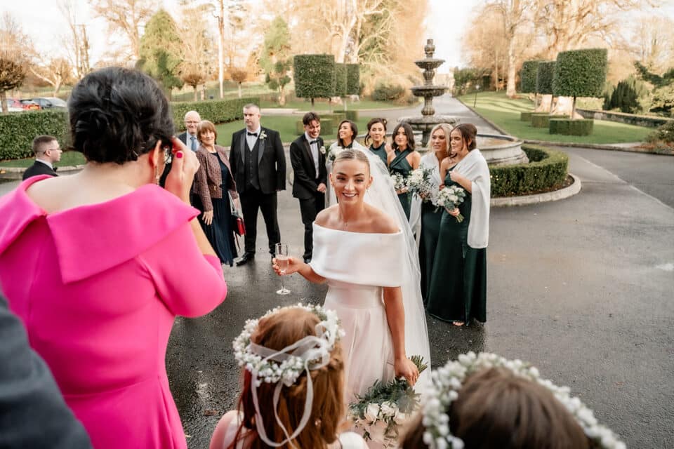 Bride in white dress with bridal party and guests at outdoor wedding reception by a fountain.