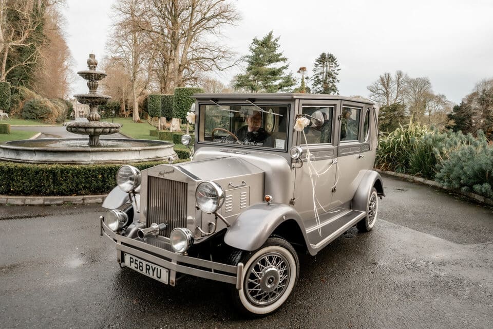 A vintage silver wedding car decorated with flowers, parked near a grand fountain and manicured gardens at Bellingham Castle.