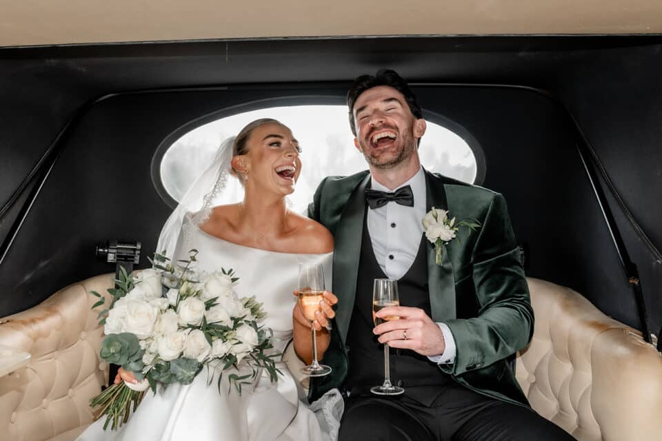 Bride and groom laughing in a car, holding champagne glasses and a bouquet.