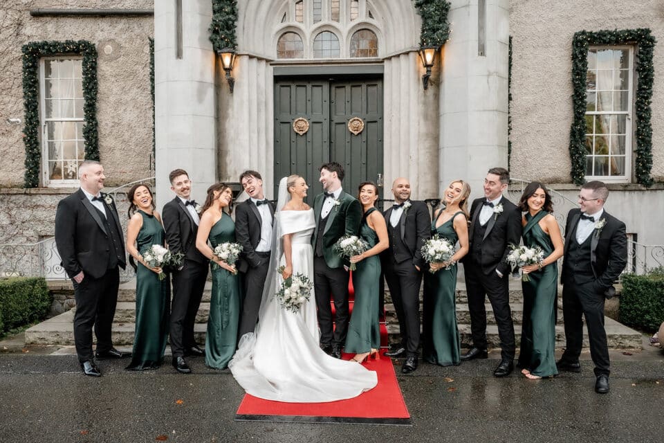 Wedding party smiling outside Bellingham Castle, bride in white gown, bridesmaids in green dresses, groomsmen in suits.