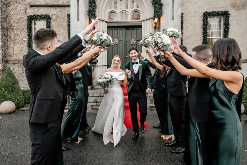 Bride and groom smiling and walking under raised flower bouquets outside a castle entrance, surrounded by bridesmaids and groomsmen.