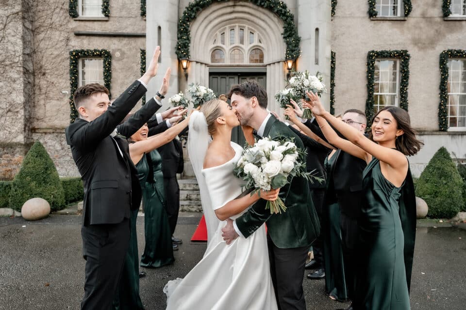 Bride and groom kissing under a raised arm archway with wedding party at Bellingham Castle.