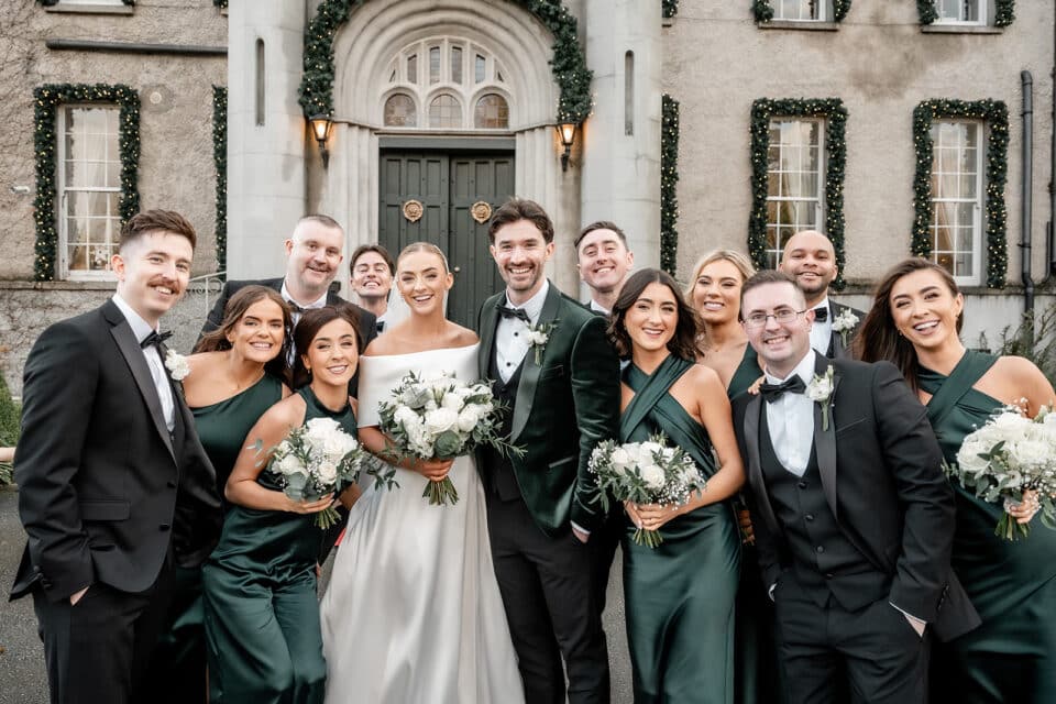 Wedding party posing in elegant attire with bouquets in front of Bellingham Castle.