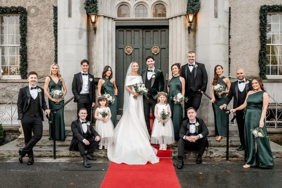 Wedding party poses on red carpet at Bellingham Castle entrance, featuring a bride, groom, bridesmaids, groomsmen, and flower girls.