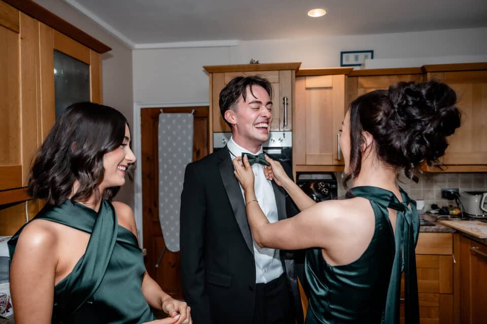 Two women assist a man with his bow tie, smiling in a warmly lit kitchen.