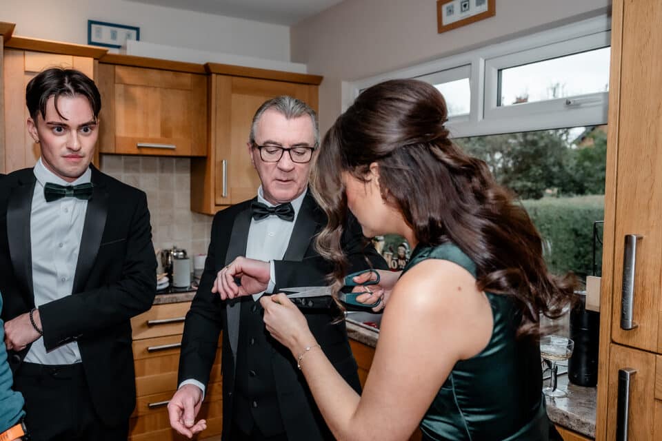 A woman cuts thread from a man's cuff while another man observes in a kitchen setting.