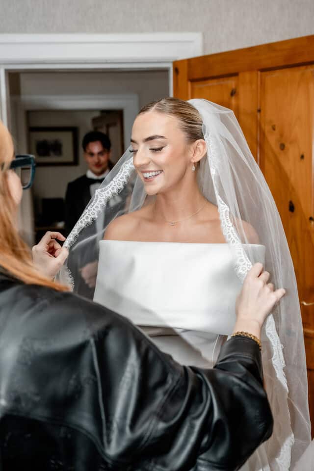 Bride smiling as her veil is adjusted, with a man in the background.