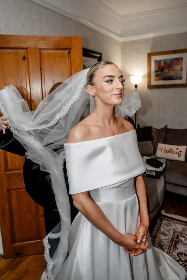 Bride in elegant white dress adjusting veil in cozy living room setting.