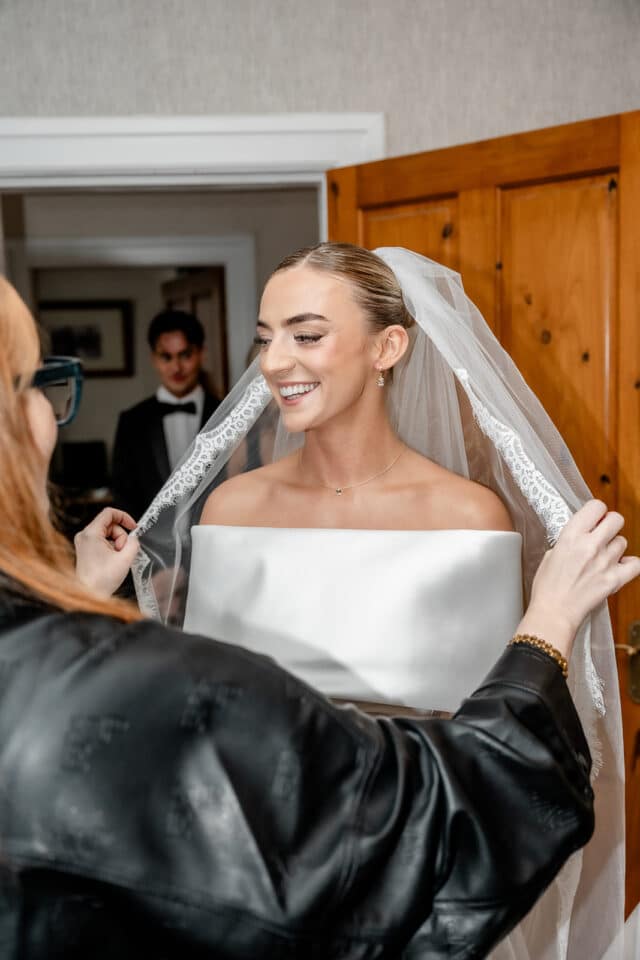 Bride smiling as she adjusts her veil, assisted by a friend, with a groom in the background at Bellingham Castle.