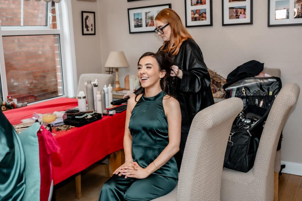 A woman in a green dress getting her hair styled by another woman in a cozy room.