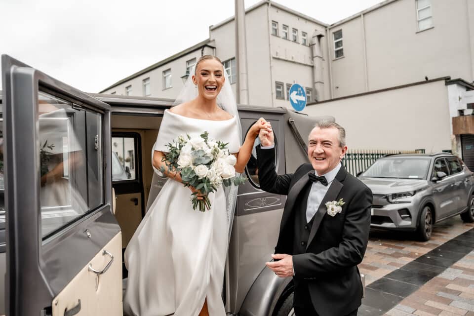 Bride stepping out of a vintage car, holding a bouquet of white roses, assisted by a suited man, both smiling.
