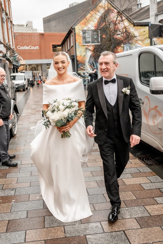 Bride in white gown holding flowers, walking with man in a tuxedo on a street near Bellingham Castle.
