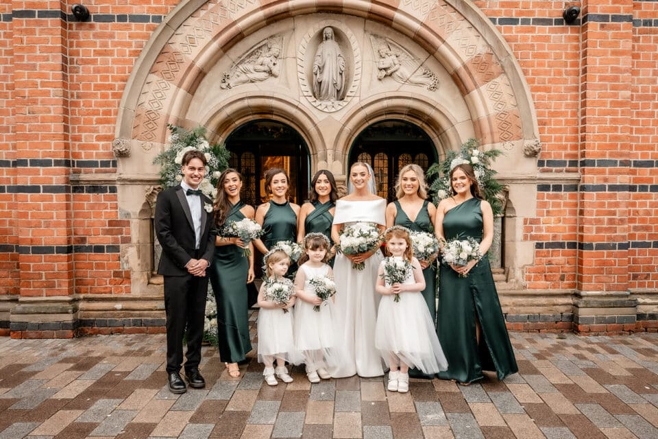 Bridal party posing in front of a brick archway at Bellingham Castle, wearing dark green dresses and holding bouquets.