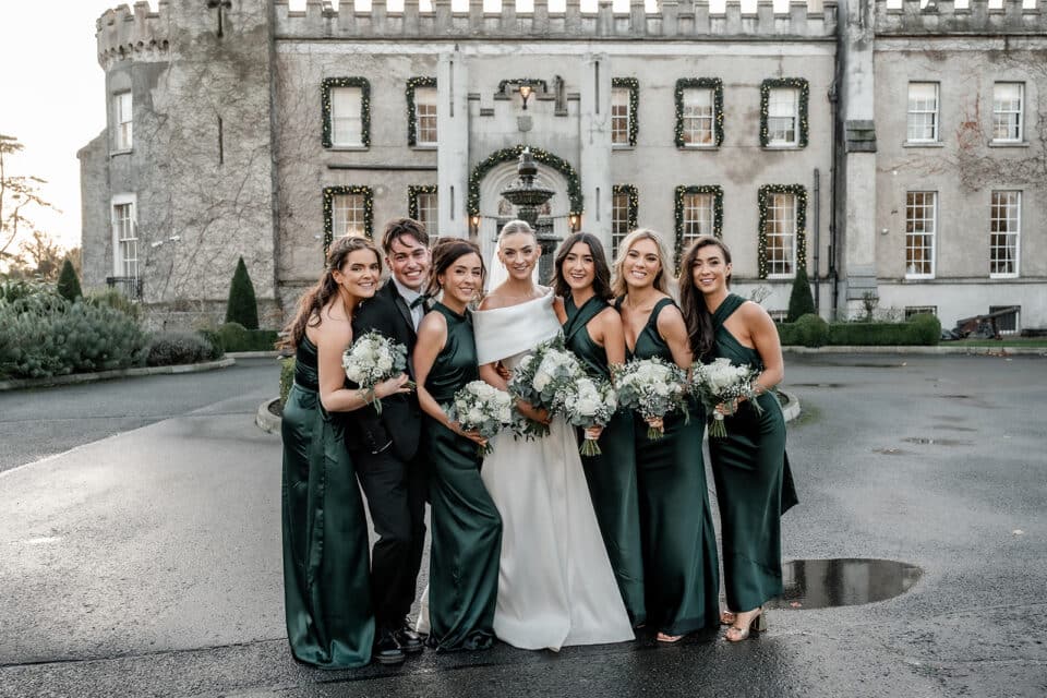 Bride, bridesmaids, and one groomsman pose with bouquets in front of Bellingham Castle, adorned in green gowns.