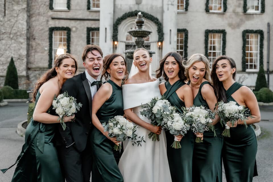 Wedding party joyfully posing at Bellingham Castle, bride in white and bridesmaids in green dresses holding bouquets.