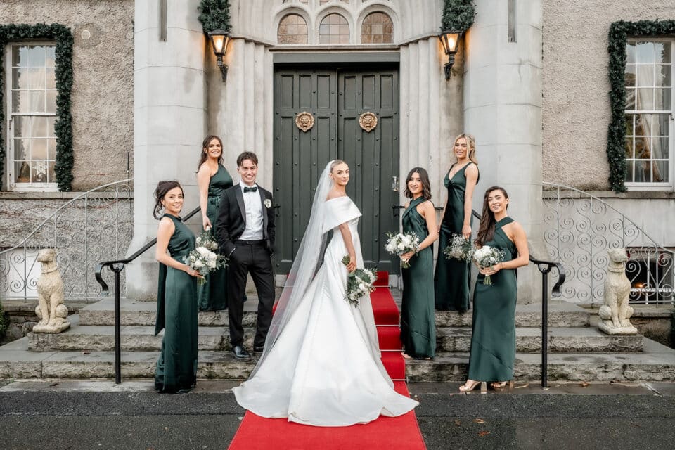 Bride, groom, and bridesmaids pose on red carpet outside Bellingham Castle, with elegant greenery decor.