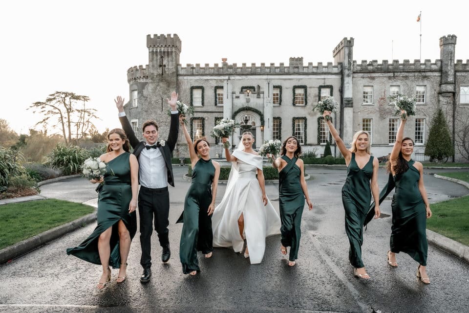 Wedding party celebrating in front of Bellingham Castle with bridesmaids in green dresses and the bride in a white gown.