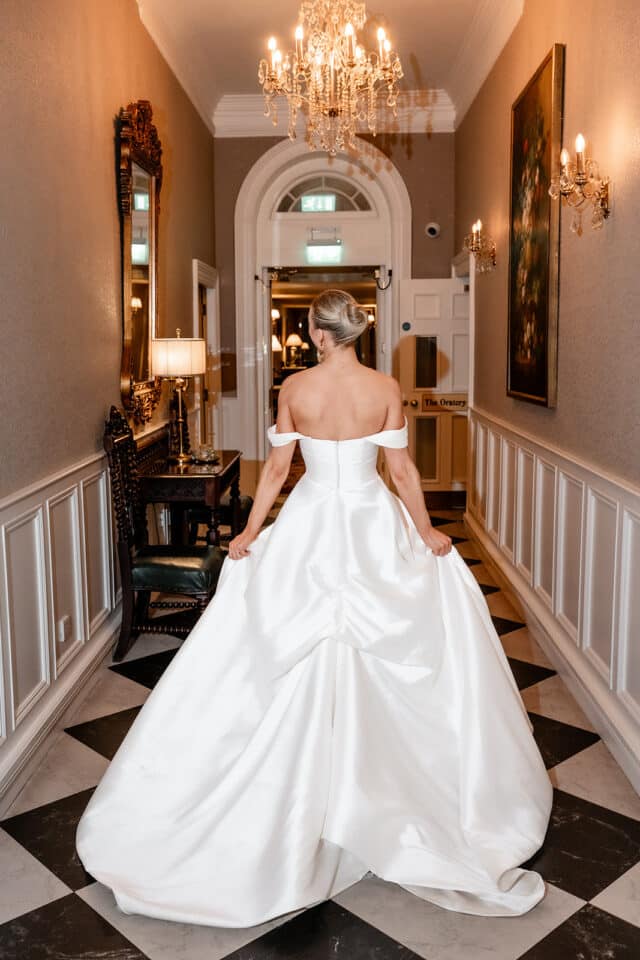 Bride in an elegant off-shoulder gown walking down a hallway with chandeliers at Bellingham Castle.