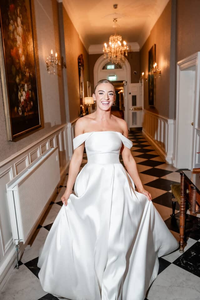 Bride in an elegant white dress smiling in a grand hallway with chandeliers.