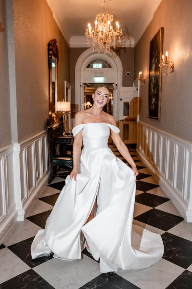 Bride in elegant off-shoulder gown smiling in ornate hallway with chandeliers at Bellingham Castle.