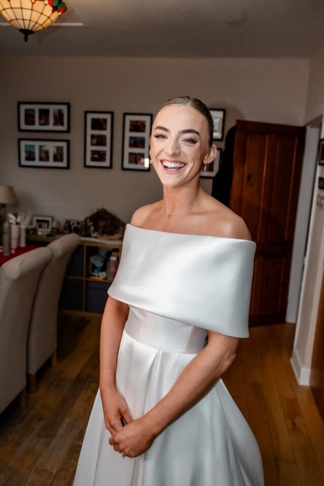 Brightly smiling bride in an elegant off-shoulder wedding dress indoors.