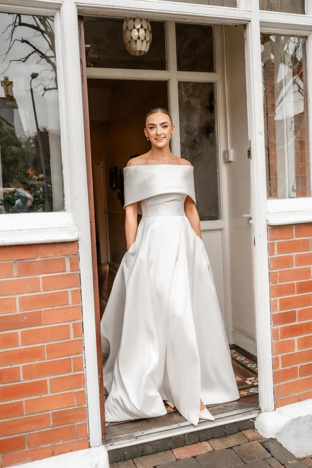Bride in elegant off-shoulder gown standing in doorway, smiling.