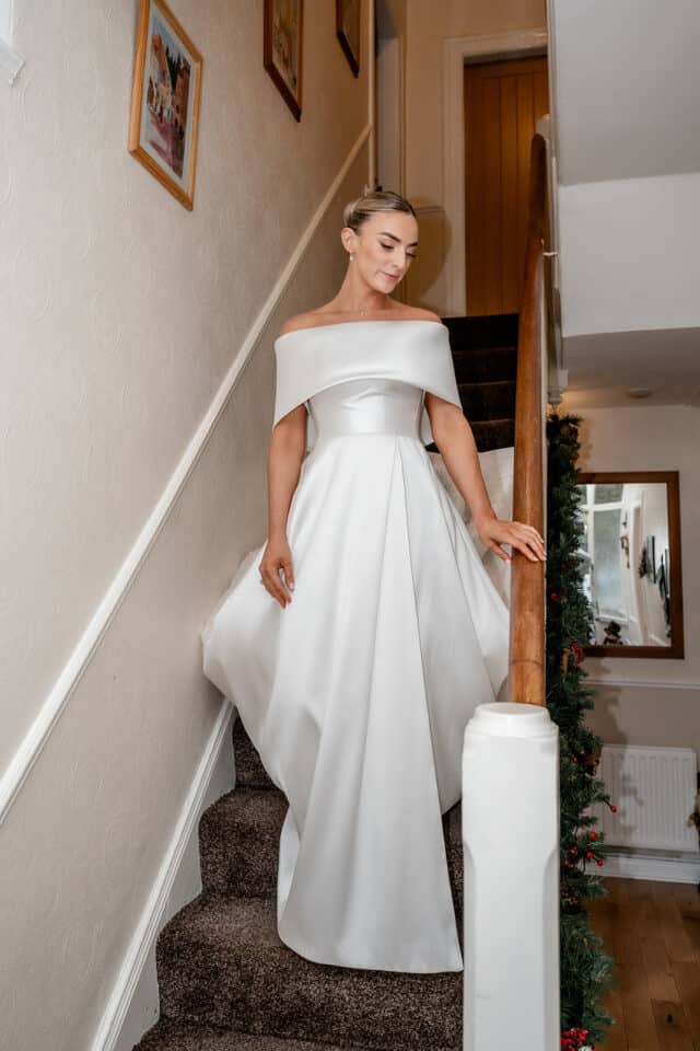 Bride in an elegant white dress descends a staircase adorned with greenery.