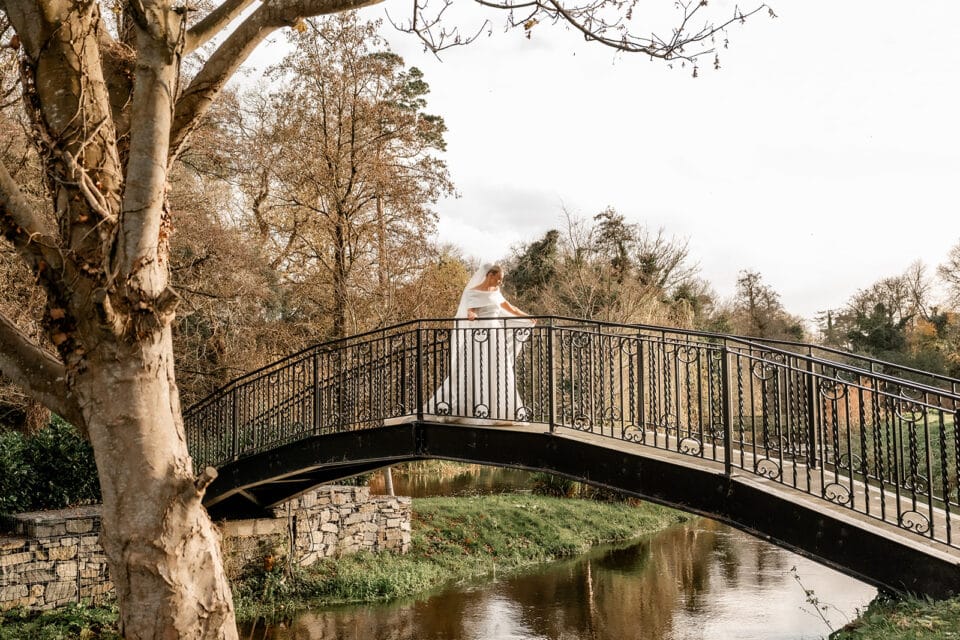 Bride in white dress standing on a bridge over a river, surrounded by autumn trees at Bellingham Castle.