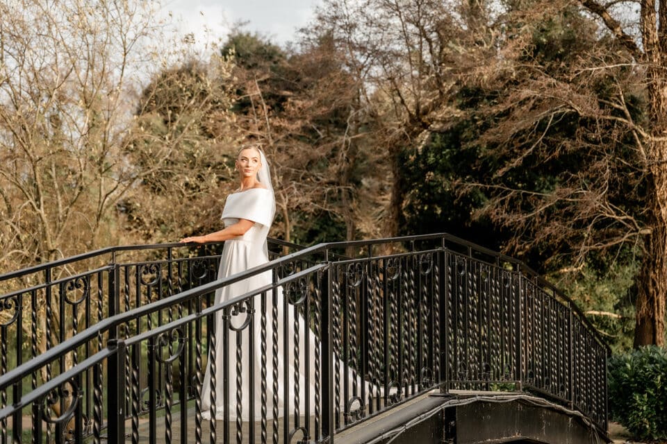 Bride in white dress stands on ornate bridge at Bellingham Castle, surrounded by trees.