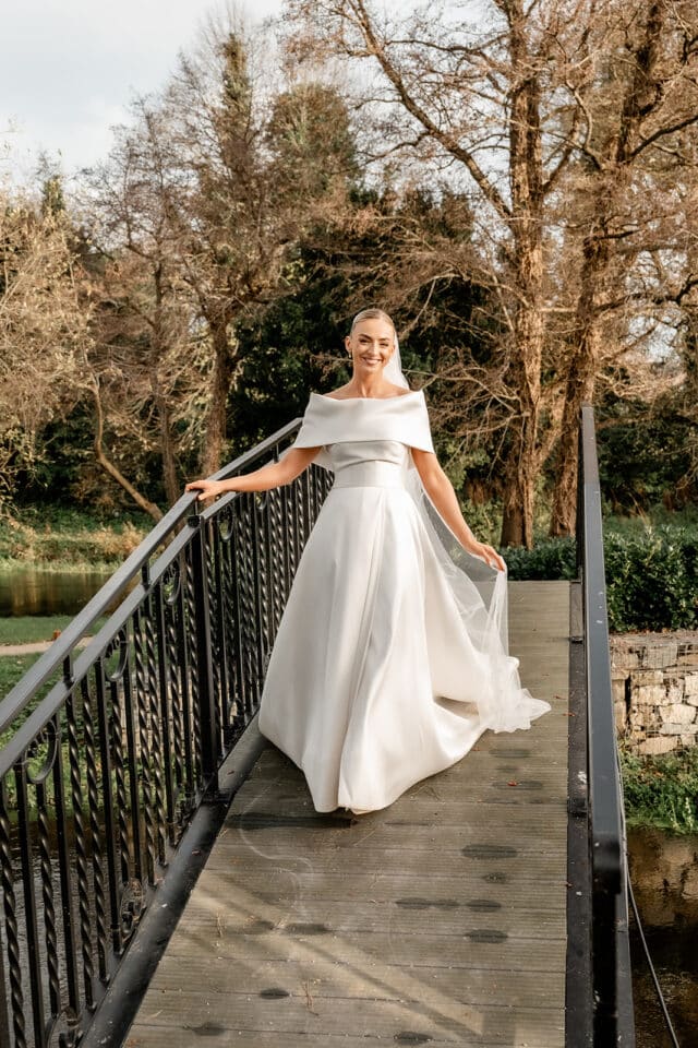 Bride in elegant white gown on a bridge at Bellingham Castle, surrounded by trees.