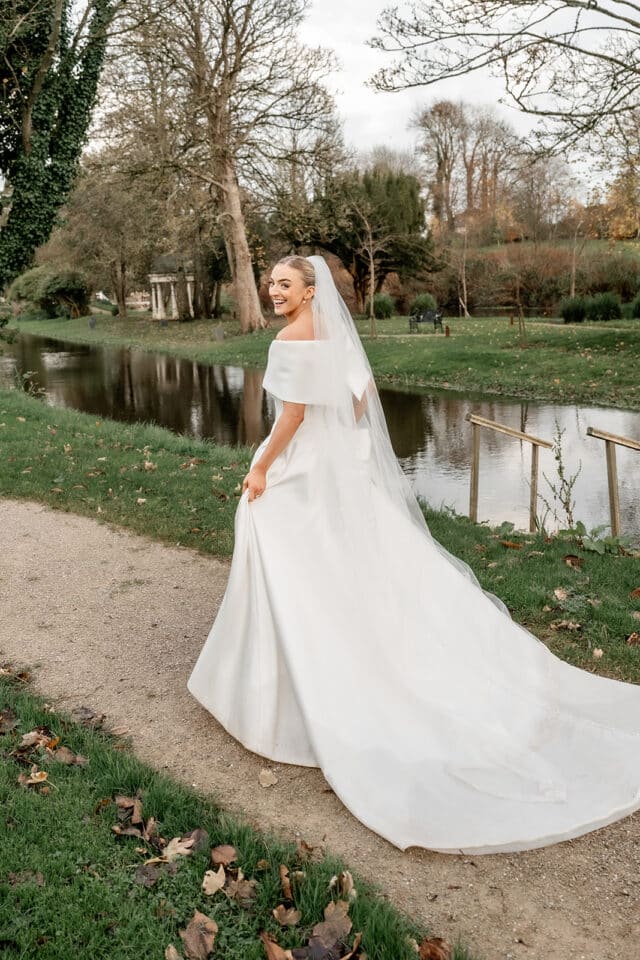 Bride in white gown and veil smiling by a pond at Bellingham Castle gardens.